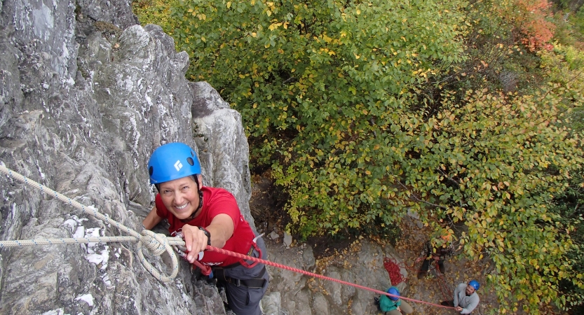 A person wearing safety gear is secured by ropes as they look up at the camera and smile while rock climbing.